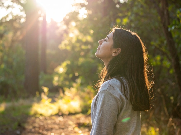 Eine Frau mit geschlossenen Augen im Wald und reckt ihren Kopf der Sonne entgegen.