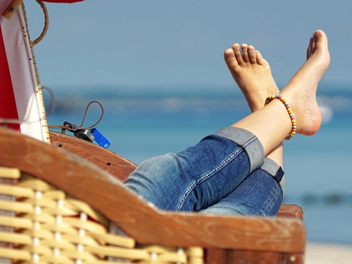 Ein Strandkorb, über dessen Lehne ausgestreckte Beine in Jeans und mit nackten Füßen ragen. Im Hintergrund Strand und Meer. 