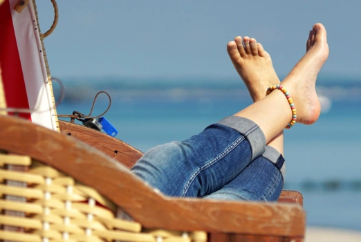Ein Strandkorb, über dessen Lehne ausgestreckte Beine in Jeans und mit nackten Füßen ragen. Im Hintergrund Strand und Meer. 