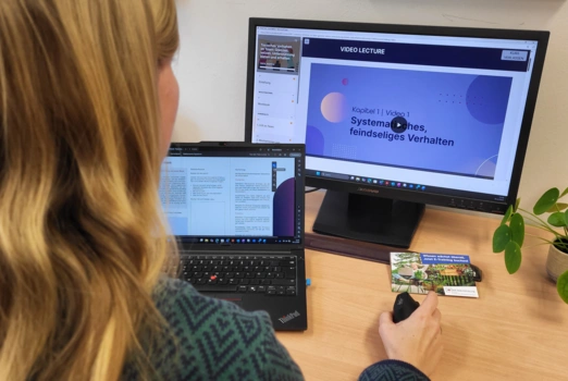 Eine Frau sitzt an einem Schreibtisch. Sie blickt auf zwei Bildschirme. Auf einem läuft ein E-Training, auf dem anderen ist ein Workbook eingeblendet.