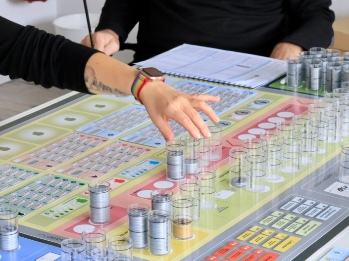 Ein Planspiel-Spielbrett mit Spielchips und Oberkörper und Hände von Spielteilnehmern, die um das Spiel herum sitzen.