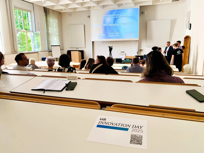 Menschen sitzen in einem Hörsaal und blicken auf ein Whiteboard. mit der Aufschrift