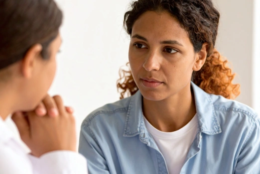 Zwei Frauen sitzen sich in einer Beratungssituation gegenüber. Eine der Frauen ist seitlich zu sehen, sie stützt das Kinn auf den Händen auf. Die andere Frau ist frontal zu sehen und blickt zu ersterer.
