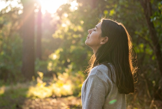 Eine Frau mit geschlossenen Augen im Wald und reckt ihren Kopf der Sonne entgegen.