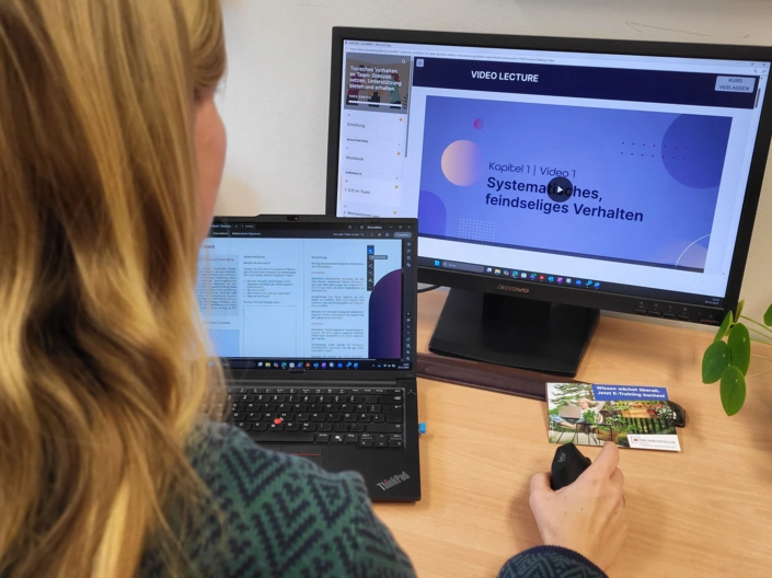Eine Frau sitzt an einem Schreibtisch. Sie blickt auf zwei Bildschirme. Auf einem läuft ein E-Training, auf dem anderen ist ein Workbook eingeblendet.