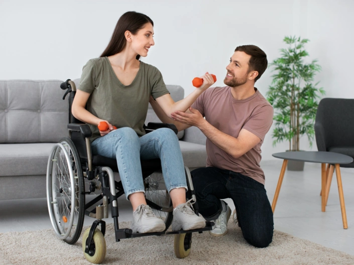 Eine Frau im Rollstuhl hat Hanteln in der Hand. Daneben ist ein Mann, der sie bei den Übungen unterstützt