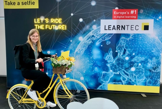 Eine langhaarige, blonde Frau in schwarzer Kleidung auf einem gelben Fahrrad vor einer Selfiewand. Die Wand trägt die Aufschrift „Learnte" und „Let's ride the future"