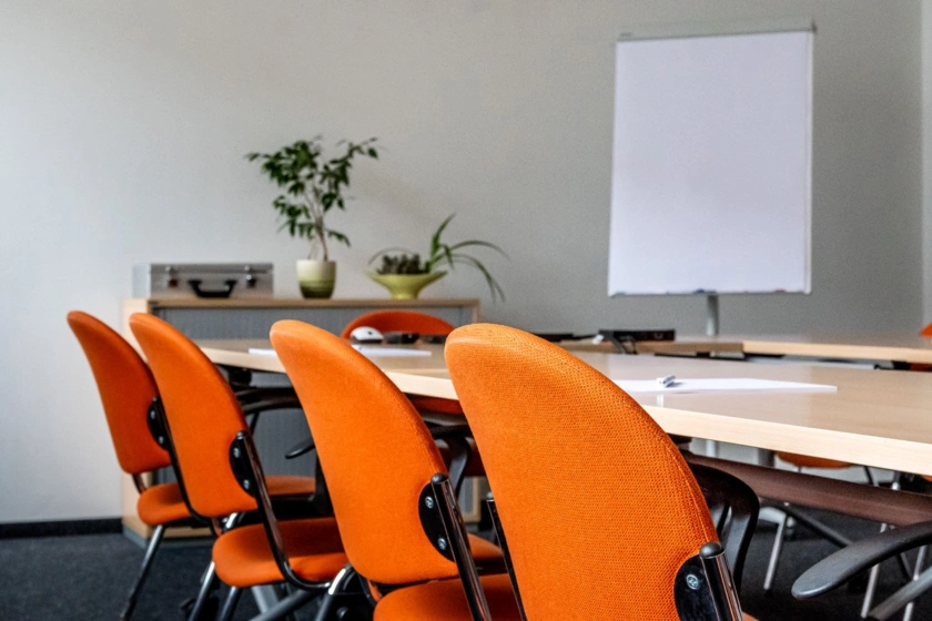 Detailansicht eines Seminarraums: Blick auf Stühle mit orangen Polstern, die an Tischen stehe. Im Hintergrund ein Flipchart und ein Sideboard mit Materialkoffer und Pflanzen.