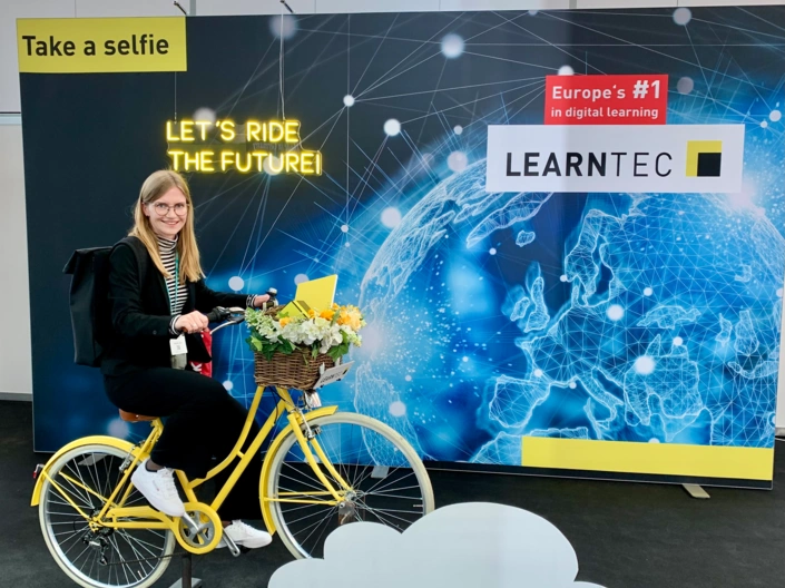 Eine langhaarige, blonde Frau in schwarzer Kleidung auf einem gelben Fahrrad vor einer Selfiewand. Die Wand trägt die Aufschrift „Learnte" und „Let's ride the future"