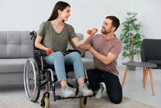 Eine Frau im Rollstuhl hat Hanteln in der Hand. Daneben ist ein Mann, der sie bei den Übungen unterstützt