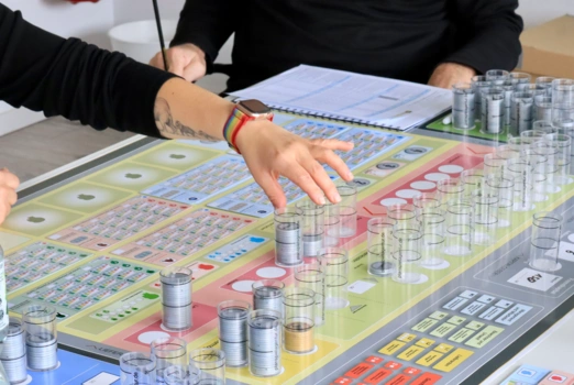 Ein Planspiel-Spielbrett mit Spielchips und Oberkörper und Hände von Spielteilnehmern, die um das Spiel herum sitzen.